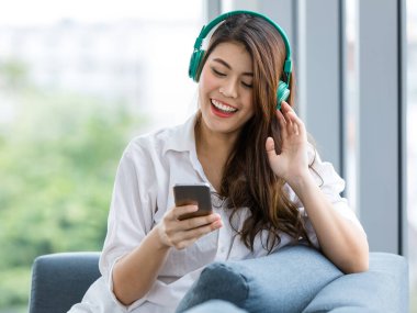 Kanepede oturan ve el rahatlatıcı şekilde basılı tutun smarthphone kulaklık ile müzik dinlemek genç güzel Asyalı kadın çekim portre kadar kapatın. Yeşil doğa ile kopya alanı sol arka plan bulanıklık.