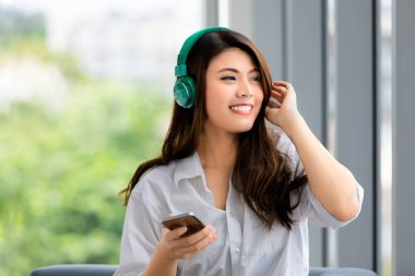 Genç, güzel Asyalı bir kadının portresini kulaklıklı, rahatlatıcı bir şekilde müziğe yakın çek ve akıllı telefonu elinde tut. Arka planda yeşil doğa bulanıklığı ve sol tarafta kopyalama alanı.