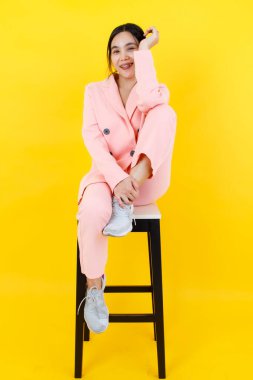 Pembe takım elbiseli sevimli Asyalı kız, dirseği dizinde sandalyeye otururken şaka yapıyor. Pastel kostümün güzel portresi, hayat dolu genç bir kadın için güzellik tasarımı..