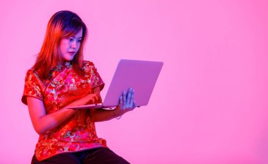 Milenyum Asyalı genç kız modelinin stüdyo fotoğrafı güzel kırmızı Çin geleneksel Cheongsam Qipao tişörtü içinde elinde dizüstü bilgisayarla pembe ışık arkaplanından alışveriş yapıyor..