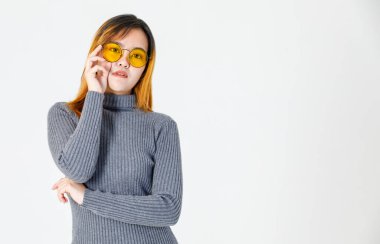 Portre yakın çekim stüdyosu gri balıkçı yaka kazaklı Asyalı genç kadın modelin el ele tutuşup moda turuncu gözlüklerine dokunurken beyaz arka planda kameraya baktığını gösteriyor..