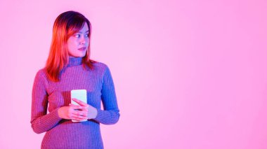 Asya 'nın modaya uygun, gri kaplumbağa yaka kazaklı, elinde dokunmatik ekranlı akıllı telefonla pembe arka plandaki boş fotokopi alanına bakarken gülümseyen kadın modelinin stüdyo fotoğrafı..