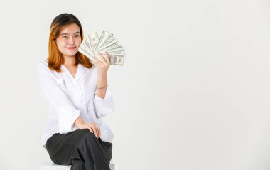 Asyalı zengin kadın iş kadını modelinin portre çekimi gündelik kıyafetlerle kameraya bakar elinde 100 dolar nakit para destesiyle beyaz arka planda durur..