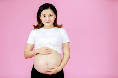 Asyalı, genç, mutlu, hamile bir kadının portre çekimi. Anne kameraya bakıyor. Açık tişört, karnına dokunuyor..