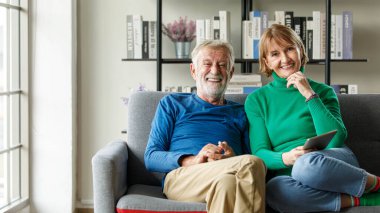 Oturma odasında kanepede birlikte otururken elinde tabletle gülümseyen ve kameraya bakan yaşlı bir adam ve olgun bir kadın.