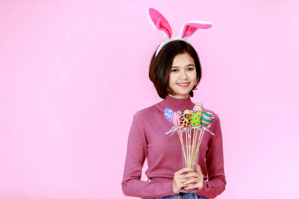 亚洲年轻快乐漂亮的短发少女模特 戴着兔子耳朵帽站在那里笑着 把传统节日彩绘的彩蛋挂在粉红的背景上 拍成电影 — 图库照片