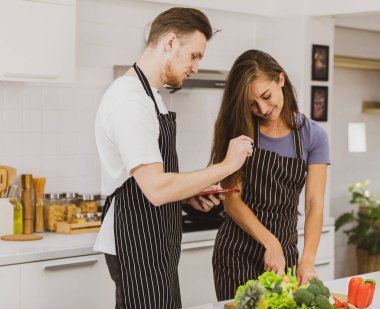 Önlüklü mutlu çift tezgahta sıcak içecekler içiyor. Mutfakta modern mutfak gereçleri ve çeşitli ürünler var.