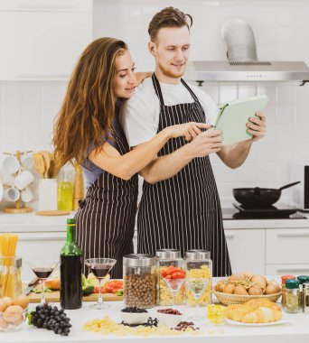 Açık modern mutfakta yemek pişirme sürecinde çeşitli ürünlerle masada dururken önlük sörfü tabletindeki memnun çift.