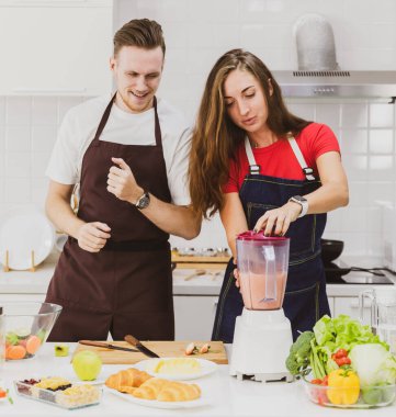 Açık modern mutfaktaki çeşitli malzemelerle blender ile ferahlatıcı smoothie yaparken önlüklü pozitif çift birlikte mutludur.