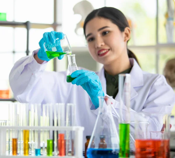 生物学の専門家は 堆積物の反応を観察するためにガラスフラスコを上げることによって青色の化学溶液液体を分析することを楽しむ アジアの女性専門家が実験室で医学研究を行う — ストック写真