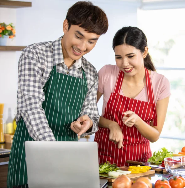 Happy Asian couples laugh and enjoy learning fun cooking course together with fingers pointing to laptop to focus on interesting recipe, instruction of training media from online cuisine at kitchen.