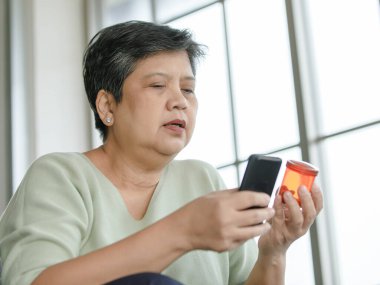 Orta yaşlı Asyalı bir kadının evde pencerenin yanında otururken ilaç şişesini incelemesi ve akıllı telefon kullanması.