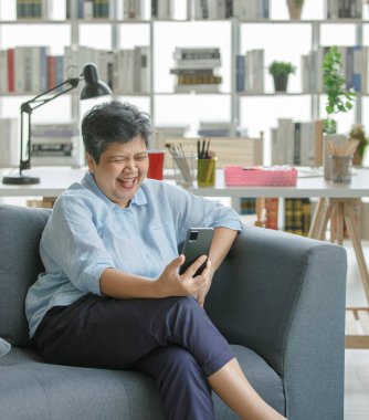 İyimser olgun Asyalı kadın gülümseyip cep telefonuyla komik videolar izlerken hafta sonu evde kanepede dinleniyor.