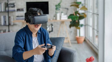 Orta yaşlı, VR kulaklıklı, gülümseyen ve video oyunu oynayan bir kadın hafta sonu evdeki oturma odasında kanepede otururken.