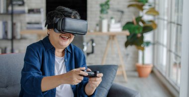 Orta yaşlı, VR kulaklıklı, gülümseyen ve video oyunu oynayan bir kadın hafta sonu evdeki oturma odasında kanepede otururken.