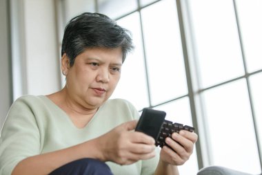 Orta yaşlı Asyalı bir kadının evde pencerenin yanında otururken ilaç şişesini incelemesi ve akıllı telefon kullanması.