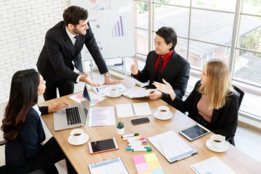 Bir grup pozitif çok ırklı iş arkadaşı, konferans odasında iş projelerini tartışırken belgeler ve aletlerle masada toplanıyor.