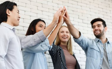 Çağdaş ışık ofisinde beyaz tuğla duvarın yanında el ele tutuşan bir grup neşeli çok ırklı iş arkadaşı.