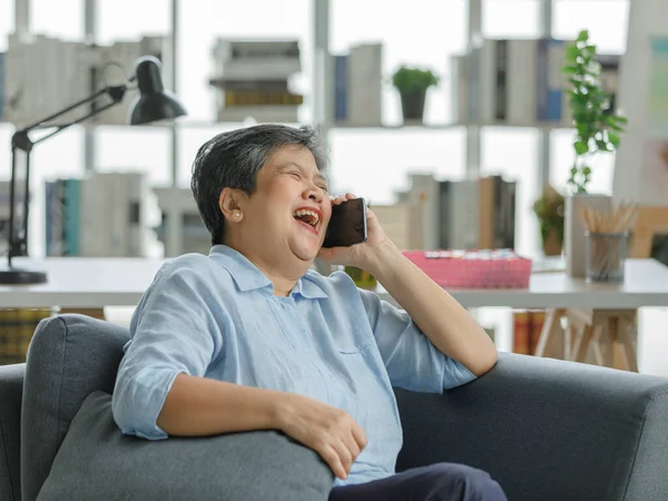 Happy mature Asian woman laughing at joke while sitting on sofa and talking on smartphone in living room at home