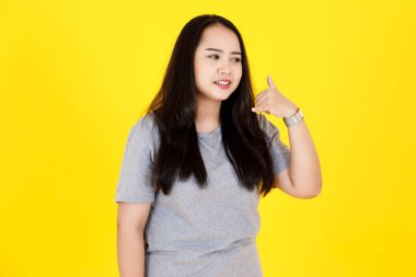 Asya 'lı genç tombul tombul, mutlu, uzun siyah saçlı, günlük gri tişörtlü, sarı arka planda telefon akıllı telefon işaretini gösteren kadın modelin stüdyo fotoğrafı..