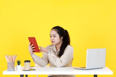 Asyalı tombul kadın sekreter, dokunmatik ekranlı tablet tutarak el sallayarak müşteriyle el sallarken, sarı arka planda video görüşmesinde müşteriyle vedalaşıyor..