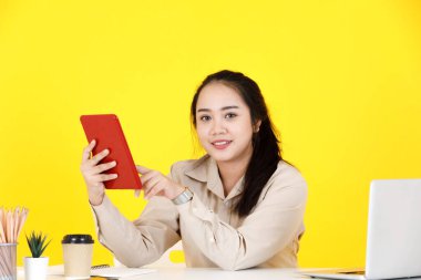 Asyalı tombul kadın sekreter, dokunmatik ekranlı tablet tutarak el sallayarak müşteriyle el sallarken, sarı arka planda video görüşmesinde müşteriyle vedalaşıyor..