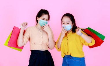 Portre stüdyosunda, pembe arka planda salgın hastalık karantinası sırasında renkli alışveriş torbaları taşıyan, yüz maskesi takan ve günlük kıyafet giyen iki Asyalı genç bayanın portresi var..