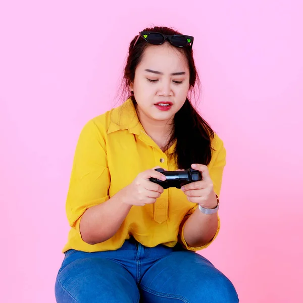 ポートレートスタジオショットのアジアの若い陽気な競争力のある女性ゲーマーでカジュアルウェアでサングラスを持って座ってジョイスティックゲームピンクの背景に — ストック写真