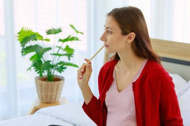 Beyaz, genç, sağlıklı, doğum öncesi hamile bir kadın. Yatak odasında ceketli, gündelik bir anne. Bebek kıyafetleri ve ekipmanları hazırlarken not defterine liste notları yazıyor..