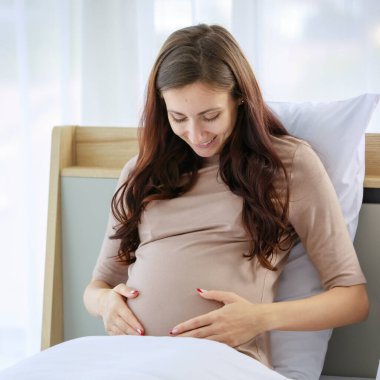 Beyaz, genç, mutlu, hamile, gündelik gömlekli bir kadın yatak odasında battaniyenin altında gülümseyen, el ele tutuşan ve karnına sevgi ve özenle dokunan bir bebek..
