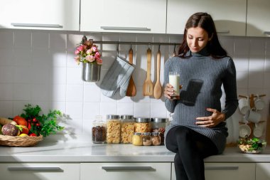 Beyaz, genç, mutlu, hamile, gündelik kıyafetlerle hamile bir kadın. Büyük göbeğin üzerinde tek başına durup sevgiyle, kahvaltıda taze süt içiyor..