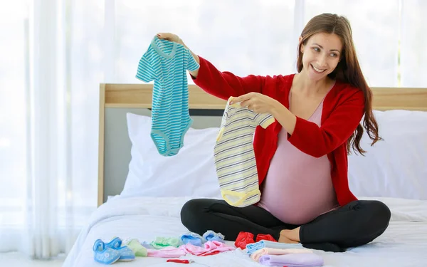 Caucasian millennial young happy female prenatal pregnant mother in casual pregnancy outfit jacket sitting smiling on bed in bedroom holding preparing choosing small cotton baby clothes in hands.