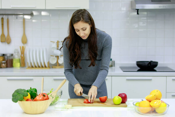 Caucasian millennial young happy sexy female prenatal pregnant mother in casual gray pregnancy dress standing smiling using knife cutting red sweet pepper on cutting board cooking vegetables salad.