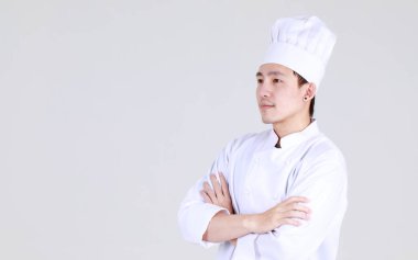 Zarif yemek pişirme üniforması giymiş deneyimli ve zeki Çinli şef. Gülümse ve kollarını kavuşturup, lüks yemekler pişirme konusunda uzman olarak mutfakta güven içinde dur.