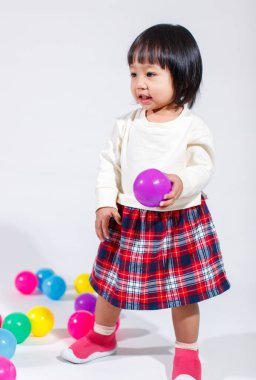 Küçük, şirin, siyah saçlı, Asyalı, ekoseli etekli ve gülümseyen beyaz arka planda tek başına renkli yuvarlak toplarla oynayan küçük bir kızın stüdyo fotoğrafı..