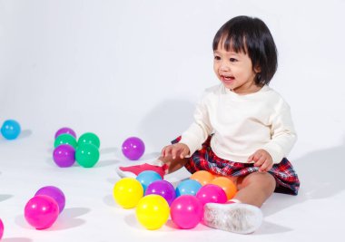 Küçük, şirin, kısa, siyah saçlı, Asyalı, ekoseli etekli, gülen, beyaz arka planda tek başına renkli yuvarlak toplarla oynayan küçük kız modelin stüdyo fotoğrafı..