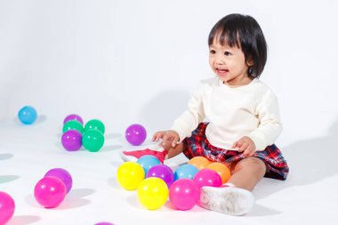Küçük, şirin, kısa, siyah saçlı, Asyalı, ekoseli etekli, gülen, beyaz arka planda tek başına renkli yuvarlak toplarla oynayan küçük kız modelin stüdyo fotoğrafı..