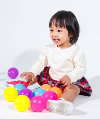 Küçük, şirin, kısa, siyah saçlı, Asyalı, ekoseli etekli, gülen, beyaz arka planda tek başına renkli yuvarlak toplarla oynayan küçük kız modelin stüdyo fotoğrafı..