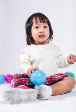 Küçük, şirin, kısa, siyah saçlı, Asyalı, ekoseli etekli, gülen, beyaz arka planda tek başına renkli yuvarlak toplarla oynayan küçük kız modelin stüdyo fotoğrafı..