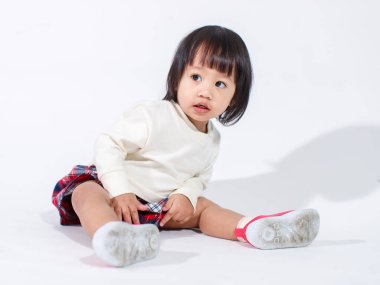 Küçük, şirin, kısa, siyah saçlı, Asyalı, ekose etekli, beyaz arka planda oturan ve gülümseyen kız çocuğunun stüdyo fotoğrafı.