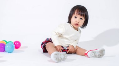 Küçük, şirin, kısa, siyah saçlı, Asyalı, ekoseli etekli, gülen, beyaz arka planda tek başına renkli yuvarlak toplarla oynayan küçük kız modelin stüdyo fotoğrafı..