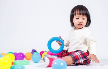 Küçük şirin Asyalı kız bebek modelin stüdyo fotoğrafı. Günlük kıyafetleriyle yerde oturmuş renkli plastik yüzüklerle oynuyorlar. İnşaat piramidi oyuncakları ve beyaz arka planda küçük toplar..