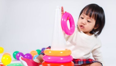 Küçük şirin Asyalı kız bebek modelin stüdyo fotoğrafı. Günlük kıyafetleriyle yerde oturmuş renkli plastik yüzüklerle oynuyorlar. İnşaat piramidi oyuncakları ve beyaz arka planda küçük toplar..