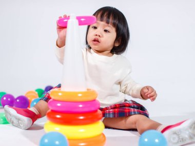 Küçük şirin Asyalı kız bebek modelin stüdyo fotoğrafı. Günlük kıyafetleriyle yerde oturmuş renkli plastik yüzüklerle oynuyorlar. İnşaat piramidi oyuncakları ve beyaz arka planda küçük toplar..