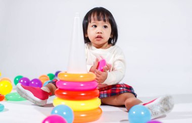 Küçük şirin Asyalı kız bebek modelin stüdyo fotoğrafı. Günlük kıyafetleriyle yerde oturmuş renkli plastik yüzüklerle oynuyorlar. İnşaat piramidi oyuncakları ve beyaz arka planda küçük toplar..