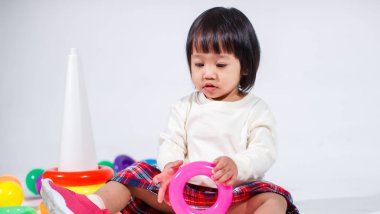 Küçük şirin Asyalı kız bebek modelin stüdyo fotoğrafı. Günlük kıyafetleriyle yerde oturmuş renkli plastik yüzüklerle oynuyorlar. İnşaat piramidi oyuncakları ve beyaz arka planda küçük toplar..