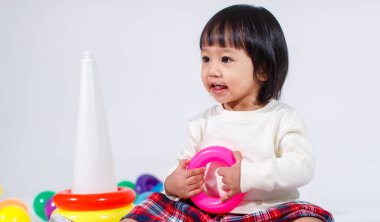 Küçük şirin Asyalı kız bebek modelin stüdyo fotoğrafı. Günlük kıyafetleriyle yerde oturmuş renkli plastik yüzüklerle oynuyorlar. İnşaat piramidi oyuncakları ve beyaz arka planda küçük toplar..