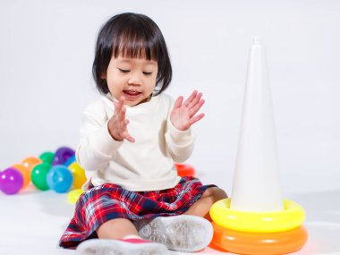 Küçük şirin Asyalı kız bebek modelin stüdyo fotoğrafı. Günlük kıyafetleriyle yerde oturmuş renkli plastik yüzüklerle oynuyorlar. İnşaat piramidi oyuncakları ve beyaz arka planda küçük toplar..