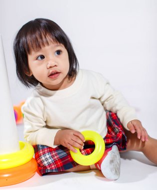 Küçük şirin Asyalı kız bebek modelin stüdyo fotoğrafı. Günlük kıyafetleriyle yerde oturmuş renkli plastik yüzüklerle oynuyorlar. İnşaat piramidi oyuncakları ve beyaz arka planda küçük toplar..