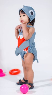 Renkli plastik toplar ve beyaz arka planda yüzük oyuncaklarıyla yerde duran şirin gri köpek balığı kostümlü Asyalı kız bebek modelinin stüdyo fotoğrafı..